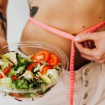 Woman holding vegetable salad and measuring waist with tape, symbolizing healthy lifestyle choices.