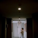 A bride stands in a dark corridor, admiring her wedding dress hanging in the light.