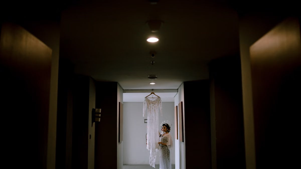 A bride stands in a dark corridor, admiring her wedding dress hanging in the light.